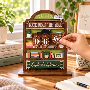 Może przedstawiać: Drewniany licznik przeczytanych książek z ciemnobrązową ramą i zielonymi półkami. Zawiera napis "BOOK READ THIS YEAR" oraz "Sophia's Library". Trzy owalne drewniane zawieszki pokazują liczbę 066. Wyświetlacz zawiera ilustracje kotów, roślin doniczkowych i książek.