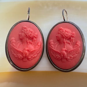 May include: A pair of oval-shaped drop earrings featuring a coral-colored cameo relief of a profile figure with an eagle. The cameos are set in a dark metallic frame and suspended from simple silver-toned metal ear wires.