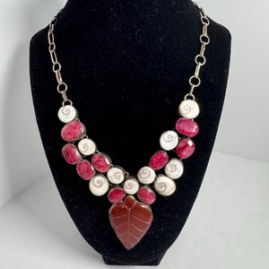 May include: A necklace featuring a central leaf-shaped reddish-brown stone pendant, surrounded by alternating oval-cut deep pink stones and circular white spiral shell beads, all set in metal and attached to a simple silver-toned metal chain link.