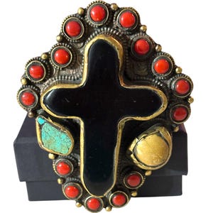May include: A large statement ring featuring a central black onyx cross set in gold-tone metal. The cross is surrounded by a border of small round red coral cabochons, a single irregular turquoise stone on the left, and a smooth tan stone on the right.