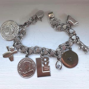May include: A silver-toned charm bracelet featuring a double-link chain with various dangling charms, including a crab, a bowling pin, a coin inscribed with MY GRADUATION DAY, a rectangular LOVE charm, a small clear stone, and a dog figure.