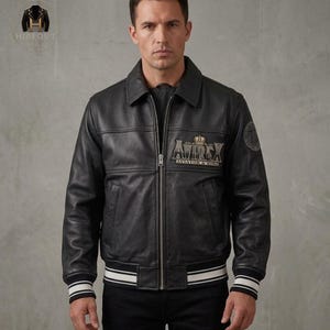 Könnte beinhalten: Eine schwarze Lederjacke mit Frontreißverschluss, klassischem Kragen und gerippten Bündchen mit weißen Streifen. Auf der Brust befindet sich ein gestickter Aufnäher mit Krone und dem Text AVIREX AVIATOR & RING sowie ein runder 75-Aufnäher am Ärmel.