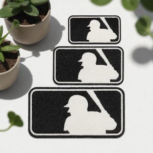 May include: Three black and white baseball patches with the MLB logo. The patches are rectangular with rounded corners and feature a batter silhouette. The patches are in different sizes.