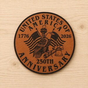 May include: A round tan leather coaster featuring a black engraved design of a bald eagle, the Statue of Liberty torch, and the Liberty Bell. The text reads UNITED STATES OF AMERICA, 1776, 2026, and 250TH ANNIVERSARY, framed by two American flags.