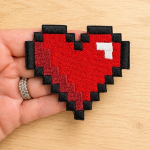 May include: A pixelated heart-shaped embroidered patch featuring a vibrant red center, a small white square highlight in the upper right corner, and a thick black border outlining the entire shape, designed in a classic 8-bit video game aesthetic.