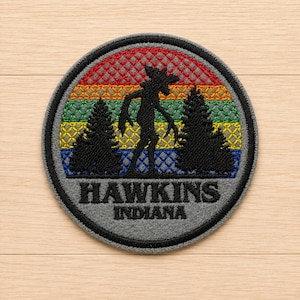 May include: A circular embroidered patch featuring a black silhouette of a creature standing between two pine trees against a background of horizontal rainbow stripes. The lower grey section displays the black text HAWKINS INDIANA.