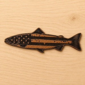 May include: A flat, fish-shaped wooden cutout featuring a laser-engraved design of the American flag. The body of the fish displays a pattern of stars on the left side and horizontal stripes across the remainder of the form in dark brown.