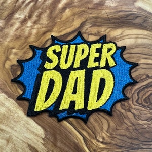 May include: A yellow and blue embroidered patch with the words "SUPER DAD" in a comic book style font.