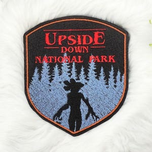 May include: A black and red embroidered patch with the text "Upside Down National Park" and a silhouette of a monster in a forest.