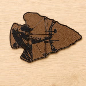 May include: A brown arrowhead-shaped leather patch featuring a black laser-engraved illustration of an archer aiming a compound bow. The design shows the archer in profile, wearing a cap, with the bow and arrow clearly detailed in black ink.