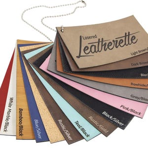 Op de afbeelding: Een waaier van kunstlederen kleurstalen, bevestigd aan een metalen kogelketting. De stalen tonen verschillende kleuren en texturen, waaronder rood, wit, blauw, bruin en zwart. De tekst "Lasered Leatherette" staat op het bovenste staal, met kleurnamen eronder.