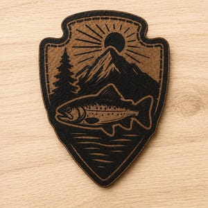 May include: A shield-shaped wooden patch featuring a laser-engraved design of a fish swimming in water below a mountain, a pine tree, and a sun. The artwork is rendered in black against a natural tan wood background with a dark border outline.
