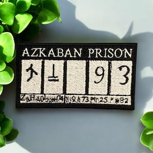 May include: Black embroidered patch with the words "AZKABAN PRISON" above a series of symbols and the numbers "193". The patch has a black border and a light beige background. Additional text is present at the bottom of the patch.