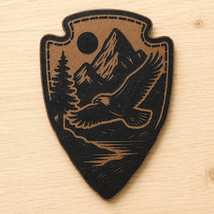 May include: A shield-shaped wooden plaque featuring a laser-engraved design of a soaring eagle, a tall pine tree, a mountain range, and a circular sun. The artwork is rendered in black against a natural tan wood background with a stitched border.