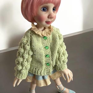 May include: A doll with pink hair and blue eyes wearing a light green knitted cardigan with green buttons. The doll is also wearing a light blue and tan dress, striped tights, and light blue shoes. The doll is a collectible item.