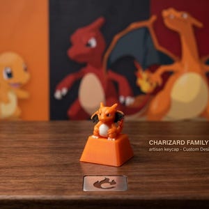 May include: An orange artisan keycap featuring a small Charizard figure with a white belly and black wings. The keycap is positioned on a wooden surface with the text CHARIZARD FAMILY artisan keycap - Custom Designed printed to the right.