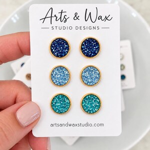 Peut inclure: Un ensemble de trois paires de boucles d'oreilles rondes sur une carte de présentation blanche marquée Arts & Wax Studio Designs. Les boucles d'oreilles ont des montures en métal doré avec des surfaces texturées et scintillantes bleu foncé, bleu clair et bleu sarcelle.