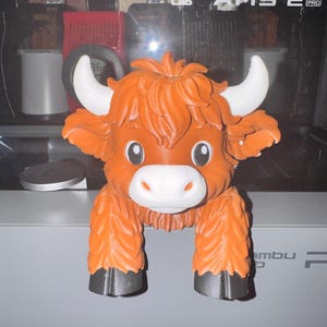 May include: A 3D printed figurine of a Highland cow with orange fur, white horns, a white muzzle, and black hooves. The cow has large, expressive eyes and a textured, shaggy appearance. The figure is designed to hang over an edge with its front legs.