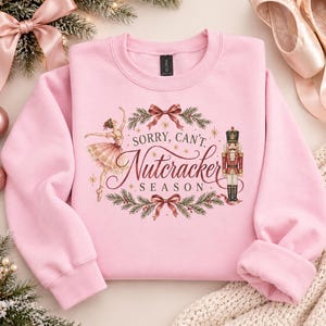May include: A light pink crewneck sweatshirt featuring a graphic print of a ballerina and a nutcracker surrounded by pine branches and bows. The text reads, Sorry, Can't. Nutcracker Season. The design is centered on the front of the garment.
