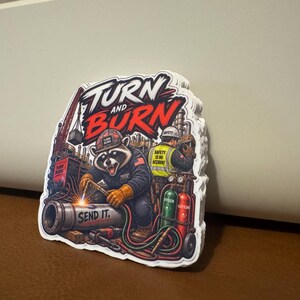 May include: A stack of die-cut stickers featuring a raccoon wearing a hard hat and welding a pipe. The graphic includes the text TURN AND BURN and SEND IT, along with industrial equipment, gas cylinders, and a worker in a high-visibility vest.
