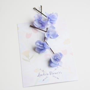Op de afbeelding: Vijf lichtblauwe stoffen bloemenhaarclips met bruine draadstengels. De haarclips zijn gerangschikt op een witte achtergrond met een bedrukt vel papier eronder. Het papier heeft een bloemenpatroon en de tekst "Linden Flowers".