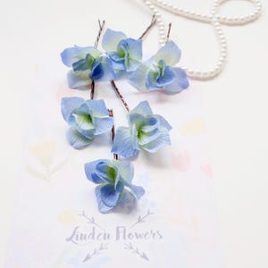 Op de afbeelding: Haarspelden met lichtblauwe hortensia bloemen en bruine stelen. Ze zijn gerangschikt op een wit oppervlak met een parelketting. De tekst "Linden Flowers" is zichtbaar in het onderste deel van de afbeelding.