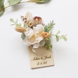 May include: A wooden sign with the text "Ailsa & Josh 2.5.25" engraved on it. The sign is decorated with a bouquet of white and beige flowers, green leaves, and brown feathers.