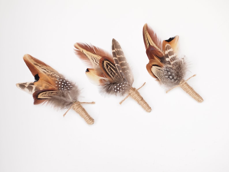 May include: Three brown and gray feather boutonnieres tied with twine. The feathers are arranged in a fan shape and have a rustic, natural look.