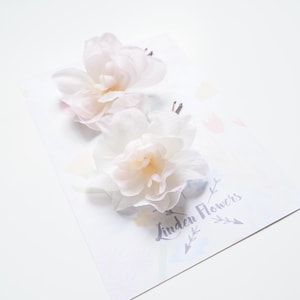 Puede incluir: Dos pinzas para el pelo con flores de tela blancas y rosa claro sobre una tarjeta blanca. Las flores tienen múltiples capas de pétalos y un aspecto suave y delicado. La tarjeta tiene el texto "Linden Flowers".