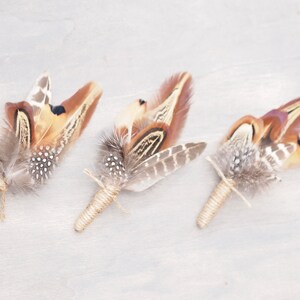 May include: Three brown and white pheasant feather boutonnieres tied with twine.