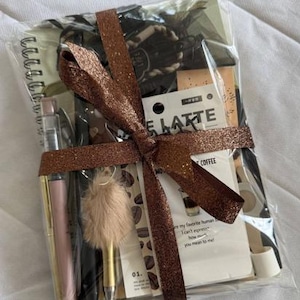 May include: A gift set wrapped in clear plastic with a brown glittery ribbon, containing a spiral-bound notebook, a pink pen, a black pen, a gold pen with a beige faux fur pom-pom topper, and a card with coffee-themed text and graphics.