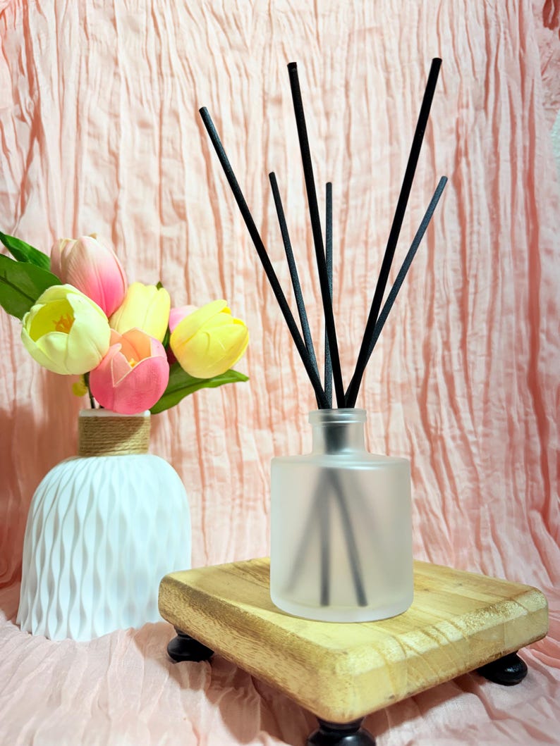 May include: A frosted glass reed diffuser bottle with five thin black reeds inserted into the neck. The bottle sits on a small square wooden riser with four dark feet. Beside it is a white vase with a textured surface containing pink and yellow tulips.