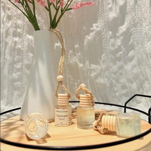 Peut inclure: Trois petits flacons diffuseurs de parfum en verre transparent avec des bouchons en bois et des cordons de suspension tressés. Une étiquette indique Modern Flame Champagne Toast 8ml. Les flacons sont disposés sur un plateau rond en bois.