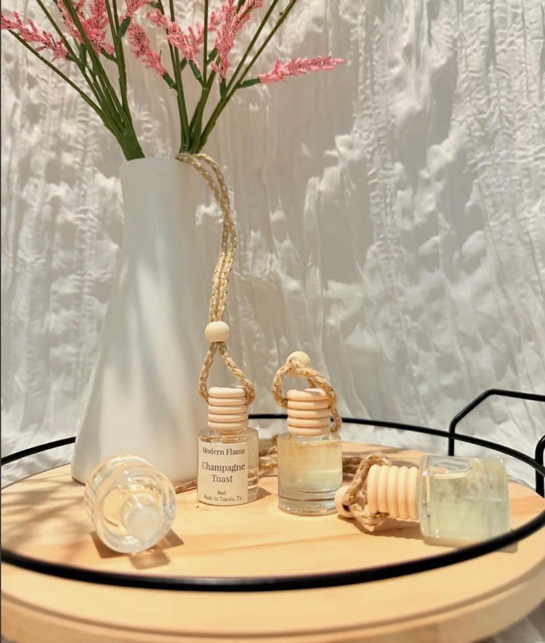 May include: Three small clear glass car air freshener bottles with wooden caps and braided hanging cords. One bottle features a label reading Modern Flame Champagne Toast 8ml. The bottles are arranged on a round wooden tray with a black metal rim.