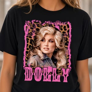 May include: A black t-shirt featuring a graphic print of a blonde person with voluminous hair. The design includes a leopard print background framed by a pink border and the word DOLLY in large pink letters at the bottom of the graphic.