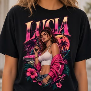 May include: A black t-shirt featuring a graphic print with the text LUCIA in large pink letters. The design includes a person holding a handgun, a pink car, stacks of cash, palm trees, a helicopter, and hibiscus flowers in a vibrant neon color palette.