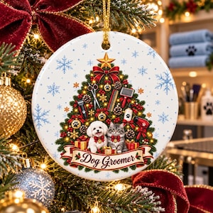 May include: A round white ceramic Christmas ornament featuring a decorated pine tree with grooming tools like scissors, combs, and clippers. A white dog and a grey tabby cat sit at the base. A red banner reads Dog Groomer.