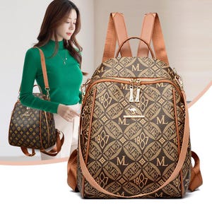 May include: A brown backpack featuring a repeating pattern of the letter M and the number 1987 in a diamond grid. The bag has tan shoulder straps, a top handle, and a gold-toned kangaroo emblem on the front pocket with two metal zipper pulls.