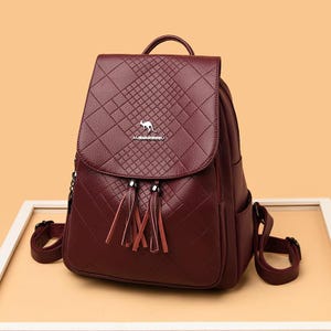 May include: A burgundy backpack with a textured, diamond-quilted pattern on the front flap. The bag features a silver kangaroo emblem, a metal logo plate, and two decorative leather tassels hanging from the centre of the front closure.
