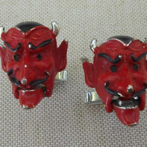May include: Pair of devil-faced cufflinks. Each cufflink features a red devil face with black eyebrows, mustache, and hair, silver horns, and a silver clasp. The devil's face has a detailed expression.