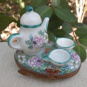 May include: A miniature porcelain tea set featuring a white teapot with a green handle and accents, and two small cups. The set is decorated with floral patterns in pink, purple, and green, and sits on a matching tray.