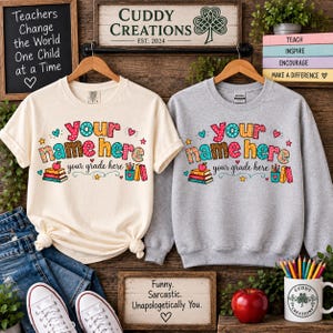 May include: A cream-coloured t-shirt and a grey sweatshirt, both featuring a colourful graphic with the text 'your name here' and 'your grade here', accompanied by illustrations of stacked books, an apple, and a pencil holder with pencils.