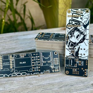May include: A black printed circuit board labeled DIYPOD SHUFFLE with gold accents, featuring an OLED screen area, button layouts, and a port for an audio jack. A second board stands vertically, displaying a black and white graphic illustration.