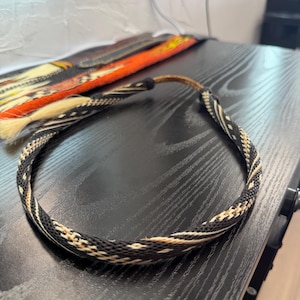 May include: A circular braided headband featuring a repeating pattern of black, white, and tan fibers. The band is constructed with a tight, woven texture and includes a smooth, brown leather-like cord section at the back for adjustable fit.