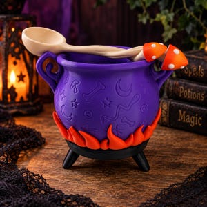 Può includere: Un calderone viola con simboli in rilievo tra cui stelle, lune e ossa, caratterizzato da una base a forma di fiamme arancioni brillanti e tre gambe nere. Un cucchiaio di legno chiaro poggia sopra, decorato con due funghi arancioni a pois bianchi.