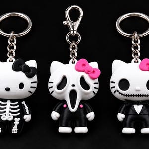 Peut inclure: Trois porte-clés personnages représentant des figures blanches ressemblant à des chats. Le premier porte un costume de squelette noir avec un nœud noir. Le deuxième a un masque fantôme avec un nœud rose. Le troisième porte un smoking noir.