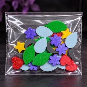Puede incluir: Una bolsa de plástico transparente llena de pequeñas formas de madera variadas, incluyendo hojas verdes, flores moradas, estrellas amarillas, corazones rojos, gotas de color azul claro y lunas crecientes de color gris oscuro.