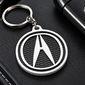 May include: A circular silver-toned keychain featuring the Acura logo in the center. The emblem consists of a stylized letter A inside a circle, set against a black honeycomb-textured background. A metal split ring is attached to the top of the charm.