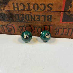 May include: Two green billiard balls with a white and black yin-yang symbol. The balls are on a white surface, with a wooden crate in the background. The crate has text that reads "Blended Scotch Whisky".