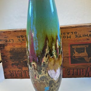 May include: A tall, cylindrical ceramic vase featuring a glossy glaze with a crackle finish. The color transitions from a vibrant turquoise at the top to deep brown and earthy tones at the base, with streaks of purple and blue dripping downward.